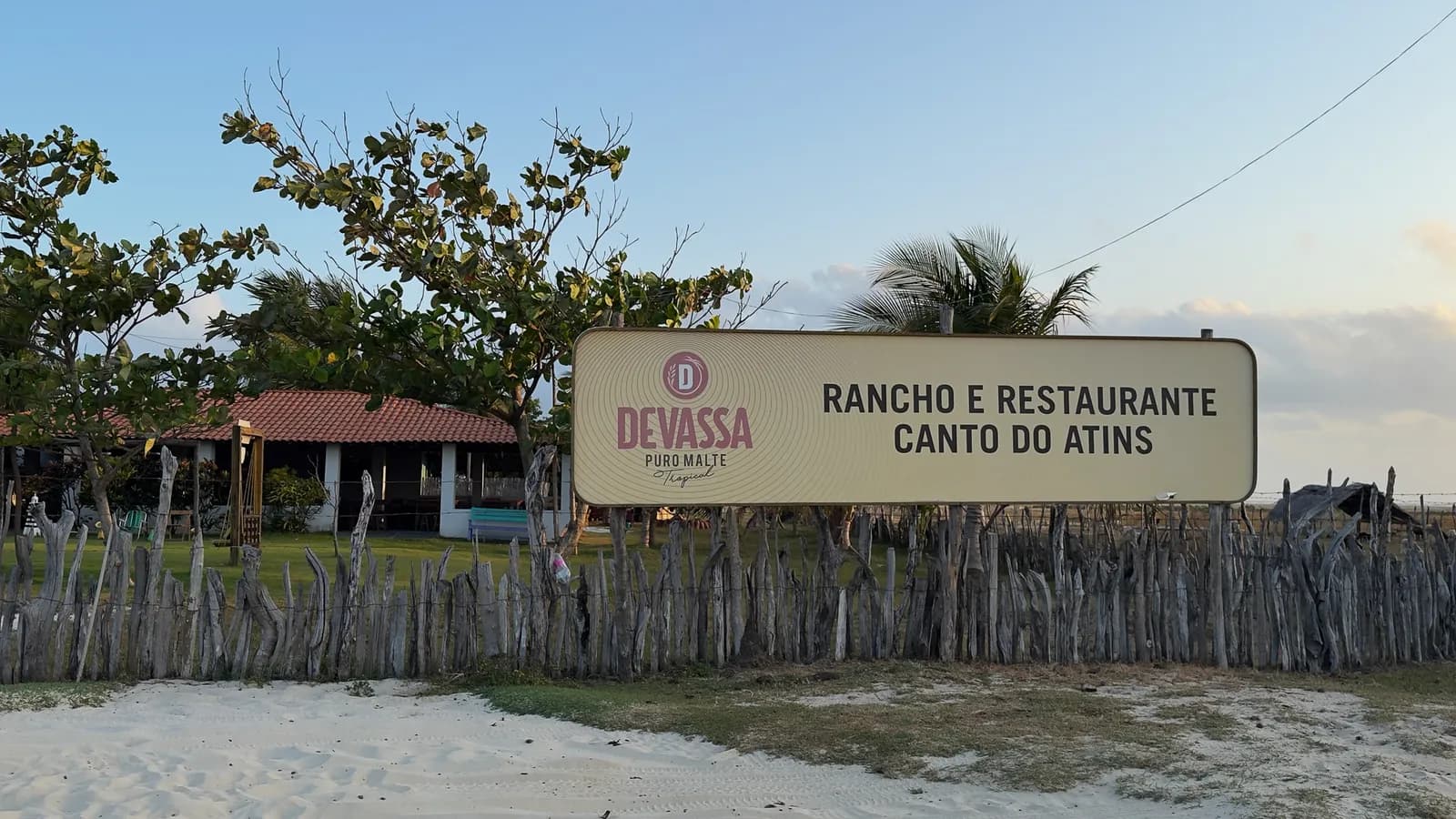 Placa na cerca de madeira com o nome Rancho e Restaurante Canto do Atins, com a areia da praia à frente